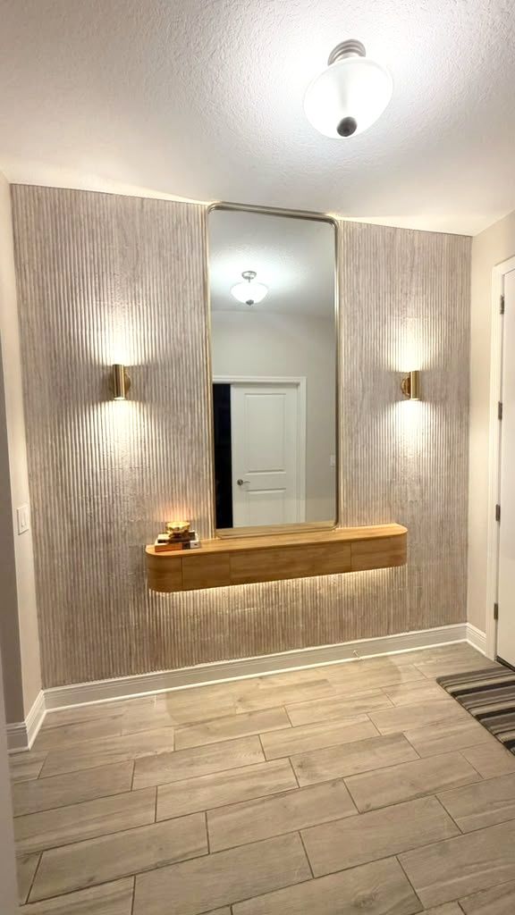 Textured accent wall with a lit floating shelf, framed mirror, and wall sconces above wood-look tile flooring.